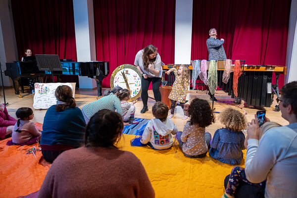 Children and parents sat on coloured rugs on the floor with three performers with instruments stood up 