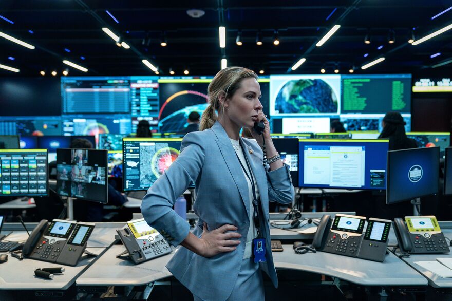 A concerned looking woman on a phone call. She stands in front of a large bank of computer monitors.
