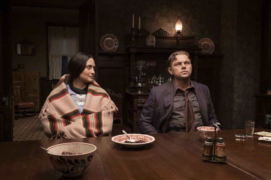 A couple sit side by side in period dress. The woman looks at the man who is staring at the ceiling. 