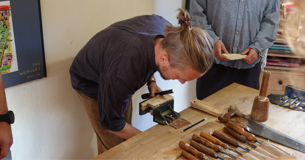 Woodcarver Tom Jennings demonstrating spoon carving at a wooden workbench fitted with hand tools and a vice.