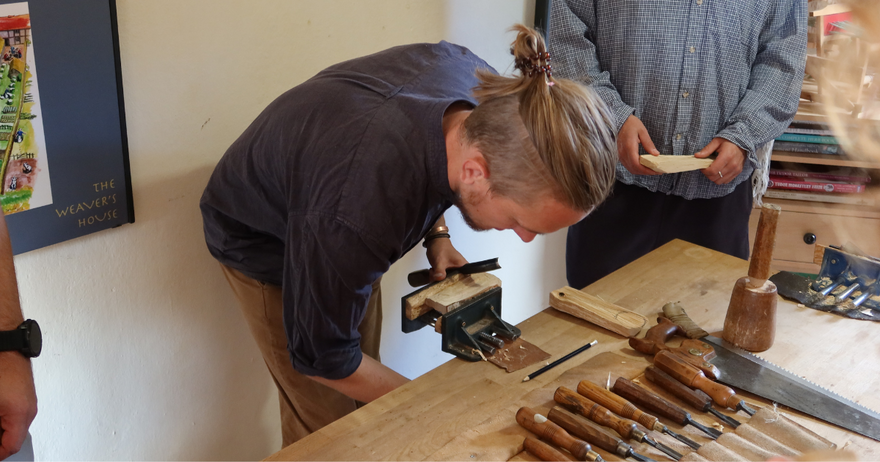 Woodcarver Tom Jennings demonstrating spoon carving at a wooden workbench fitted with hand tools and a vice.