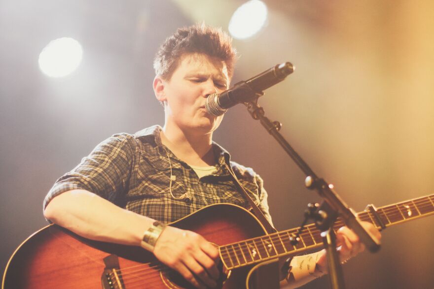 Grace Petrie sings on stage, while playing an acoustic guitar under warm, hazy spotlights.