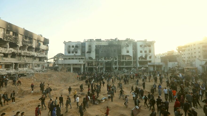 A large group of people stand amongst the ruins of destroyed buildings. 