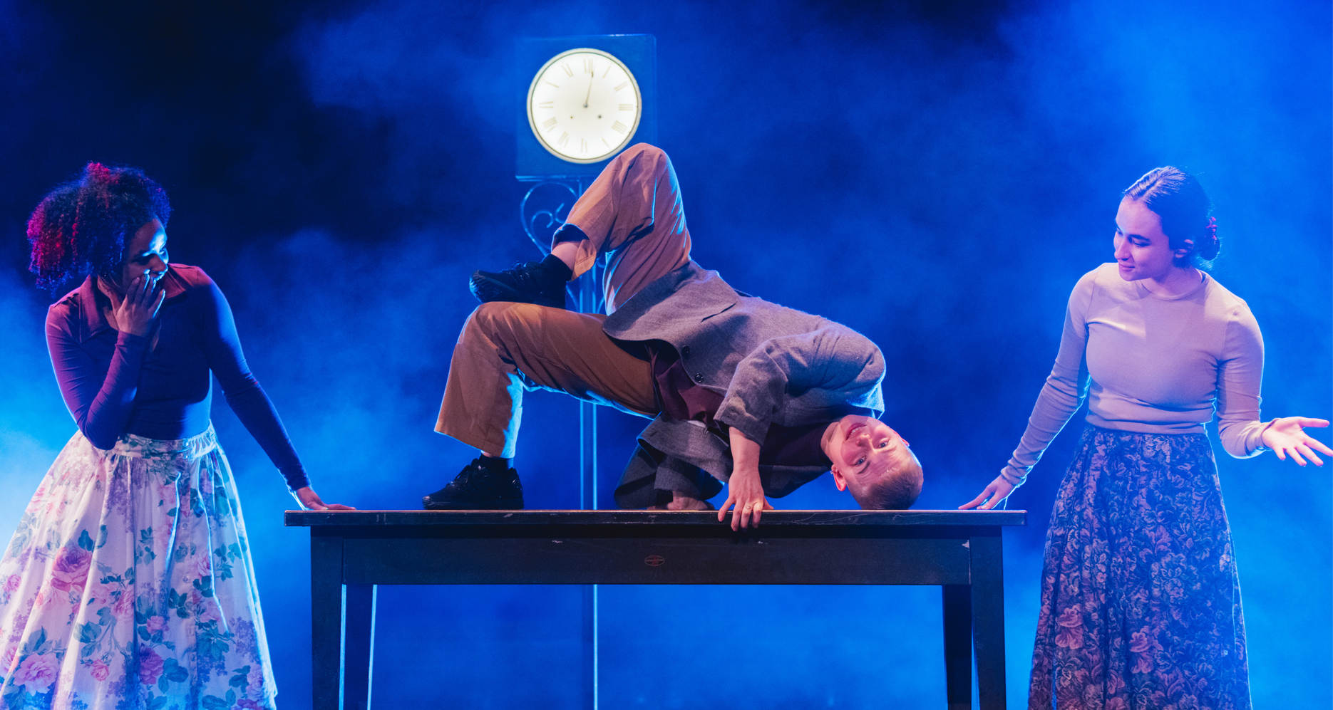 An acrobat balances upside down on a wooden table between two onlooking performers under atmospheric blue lighting.