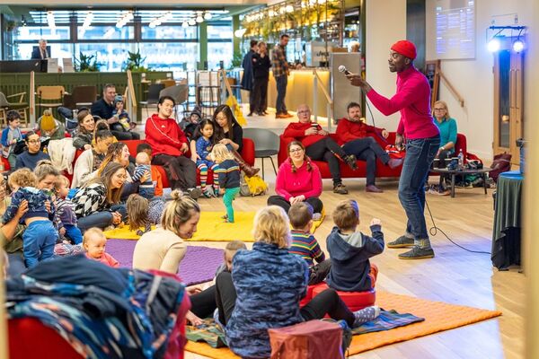 Man stood with microphone in hand with families sat on matts and sofas in front of him