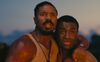 Two fearful men embrace as they watch a fire blaze. 