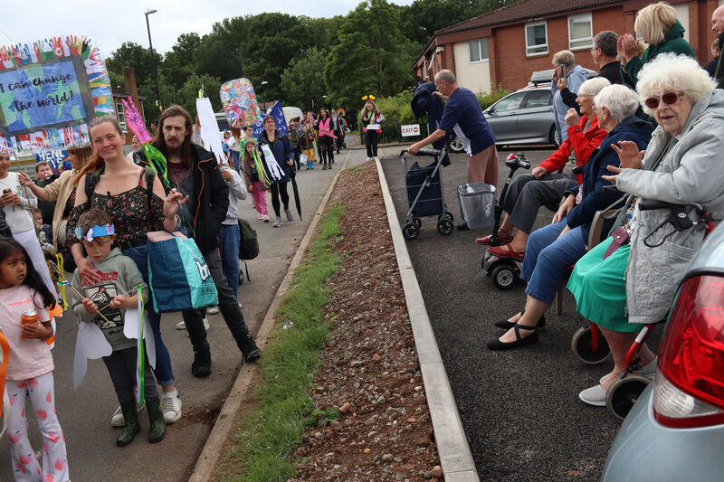 Parade participants walk down a residential street, with one carrying a large sign decorated with world flags that reads 'I can change the world!'