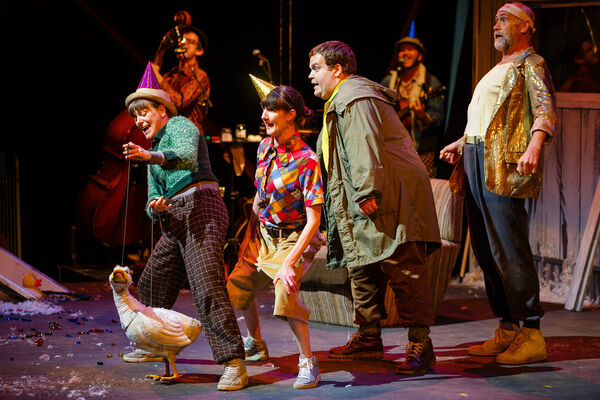 Four performers, two female and two male, in colourful clothes, stand in a line behind a puppet of a white duck