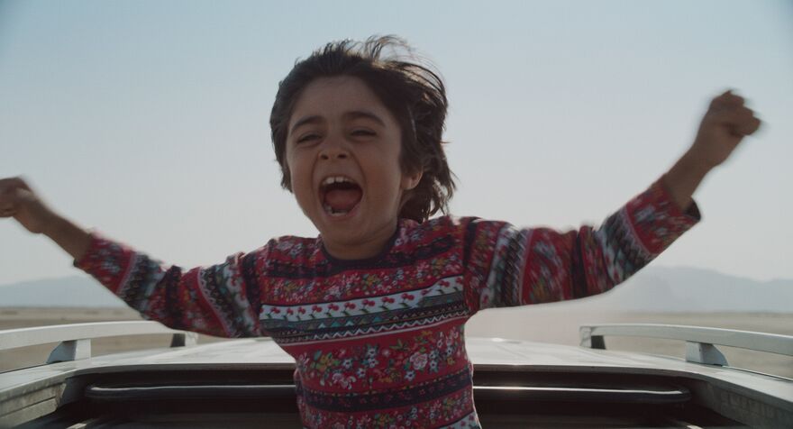A young boy stands up through a car sun roof with his arms out and an expression of joy on his face. 