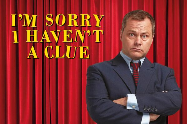 Jack Dee stood with his arms crossed in front of a red theatre curtain with 'Im Sorry I Haven't A Clue' in yellow text to the left