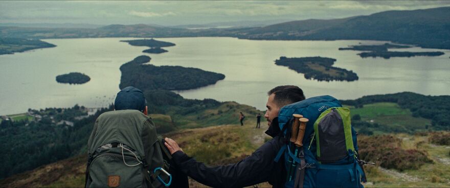 Two men sit together on a grassy hillside overlooking a lake. One man places his hand on the other's shoulder. 