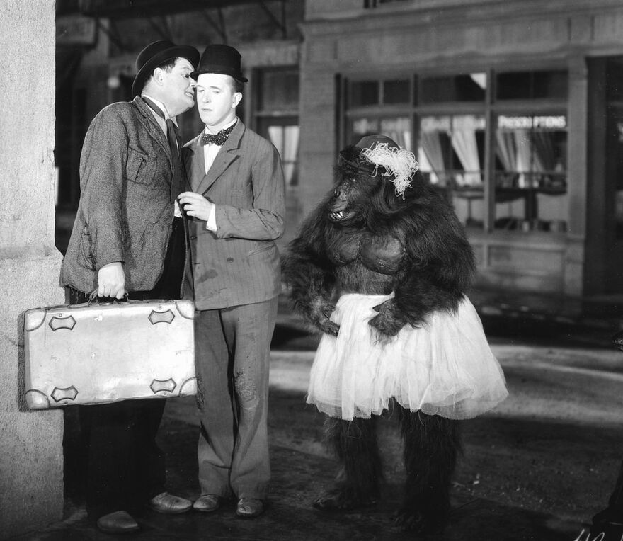 A black and white image of Stan Laurel and Oliver Hardy standing next to a gorilla in a tutu. 