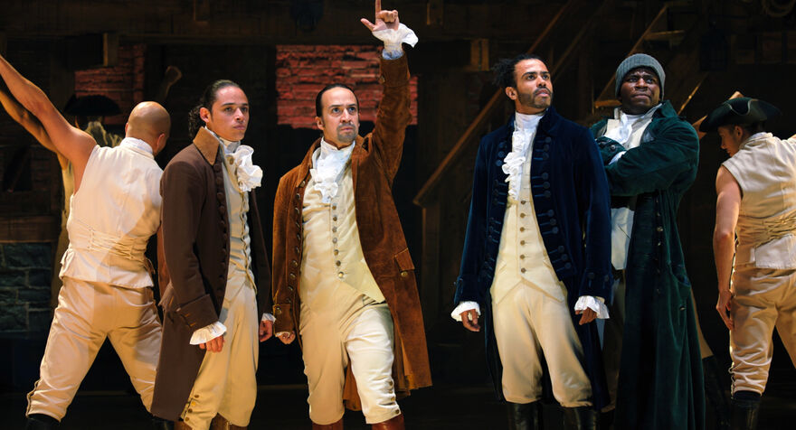 Four men in period costume stand on stage. Lin-Manuel Miranda as Alexander Hamilton holds his hand in the air.