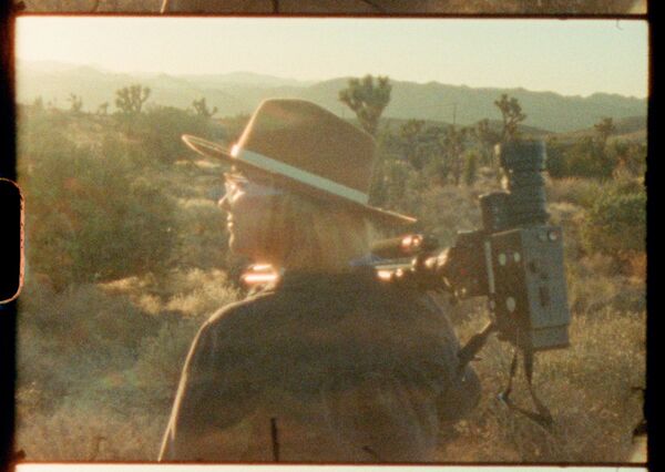 A grainy film cell of a woman in a wide brimmed hat stands amonst greenery with a camera resting on her shoulder. A grainy film cell of a woman in a wide brimmed hat stands amonst greenery with a camera resting on her shoulder.