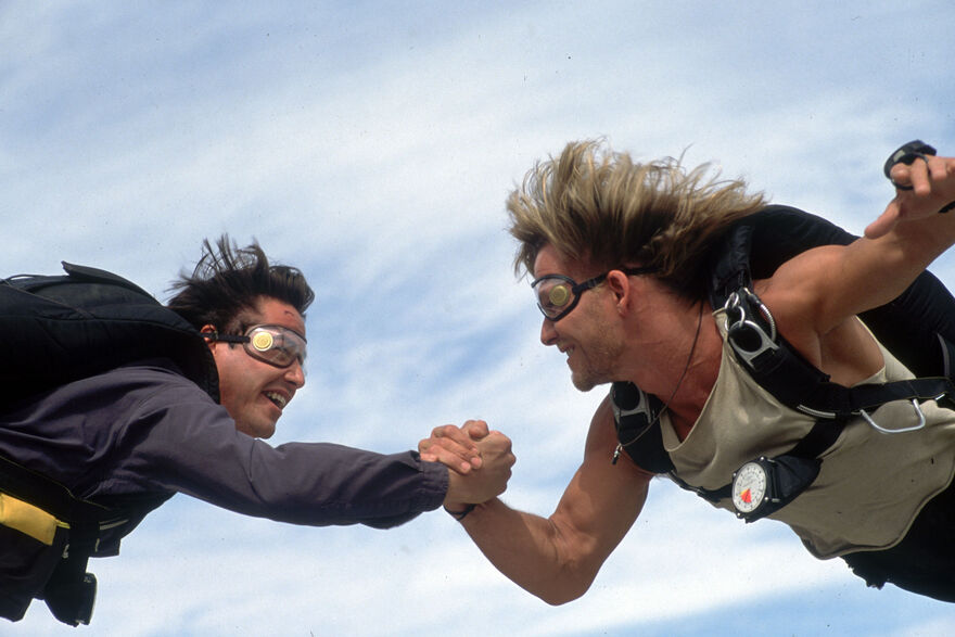 Two men hold hands as they sky dive