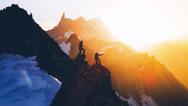 Mountains with the sun rising from behind with two people stood on the peak 