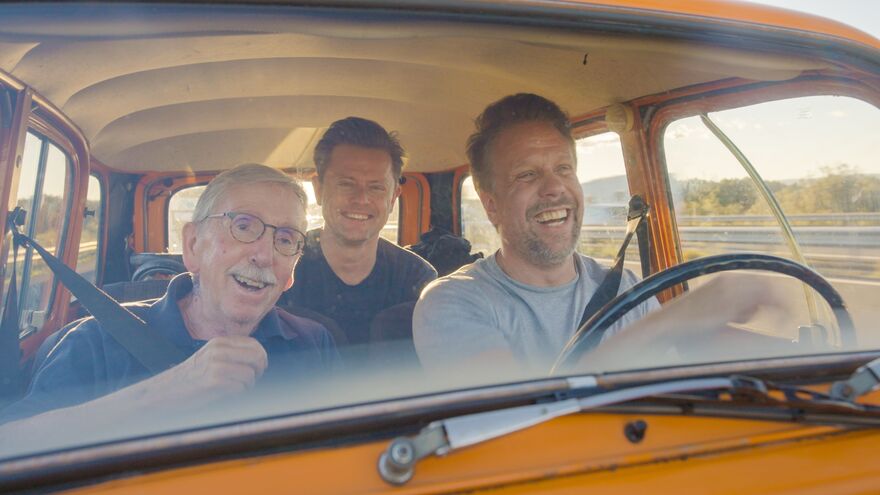 Three men sit together smiling in a yellow car.