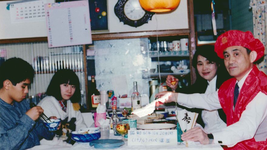 A family sit together at a dinner table looking to camera. 