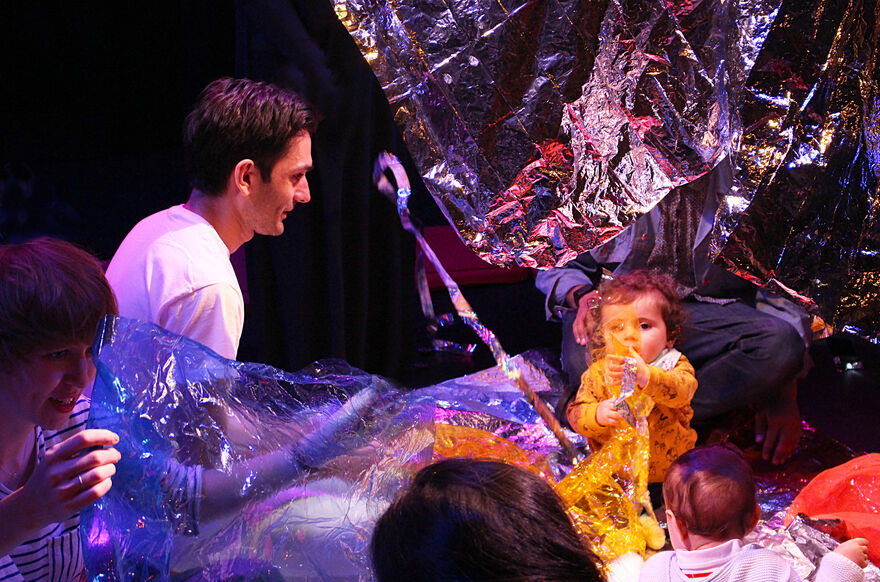 Daniel Naddafy wearing white and sitting amonst shiny materials with a number of babies playing too
