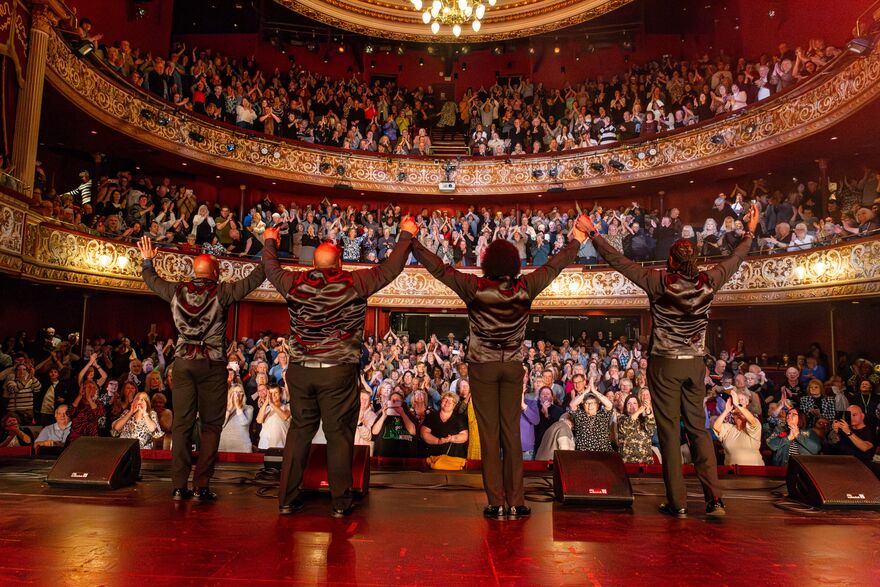 The back of four people on stage with their arms in the air in front of a crowd