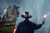 A man holds up a flare in the pouring rain as a T-Rex walks toward him. 