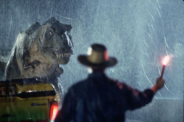 A man holds up a flare in the pouring rain as a T-Rex walks toward him. 
