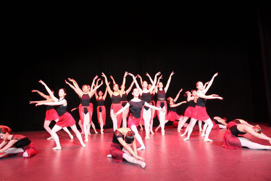A group of dancers in black tops and red skirts performing gracefully on a lit stage against a dark backdrop.