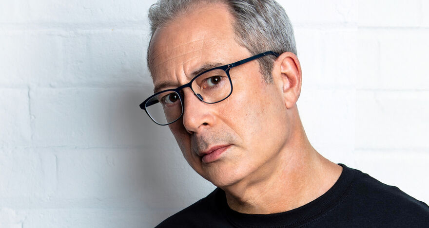 Ben Elton wearing glasses and standing in front of a white brick wall