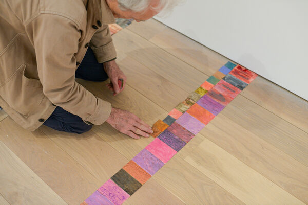 A high-angle shot of artist Martin Green kneeling on a light wooden floor and carefully arranging a long, single line of small, multi-coloured rectangular collage tiles.