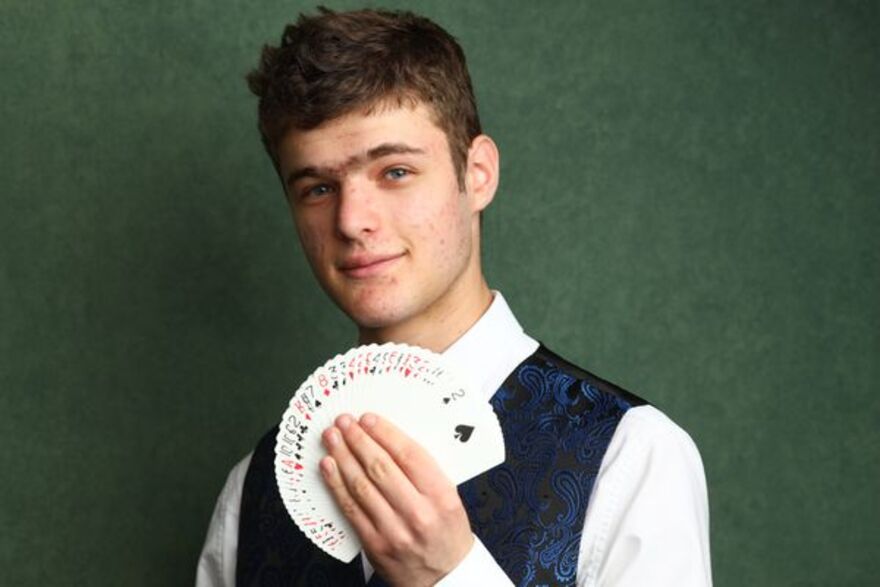 Magician Joseph smiling and holding a fanned deck of playing cards, wearing a white shirt and a blue patterned waistcoat.