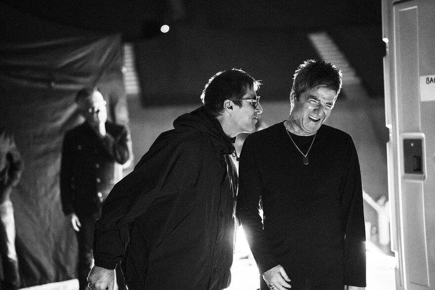 A black and white photo of Noel and Liam Gallagher laughing together backstage. 