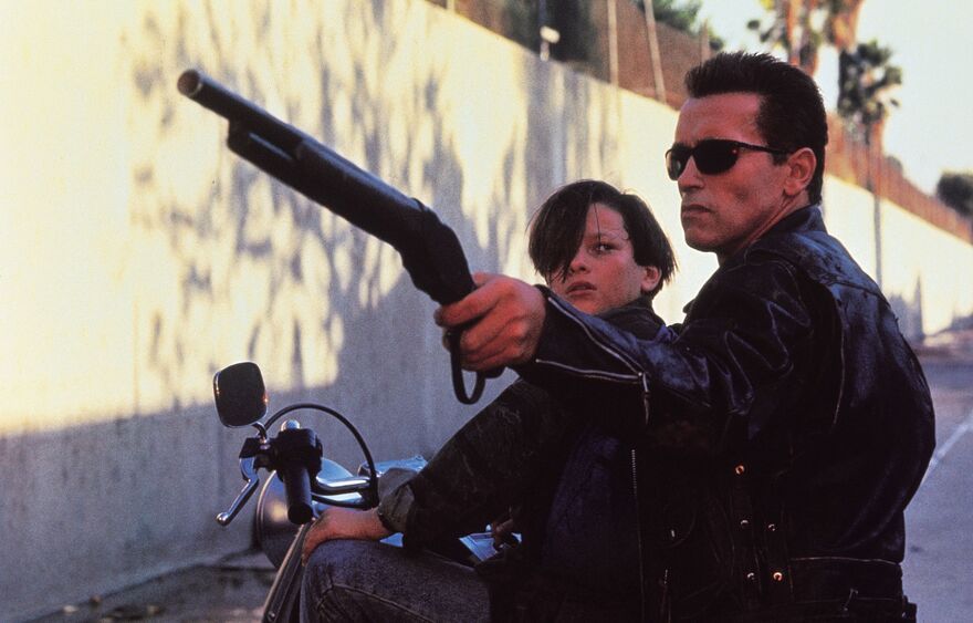 A teenage boy and a human-looking cyborg sit together on a motorcycle. The cyborg aims a rifle. 