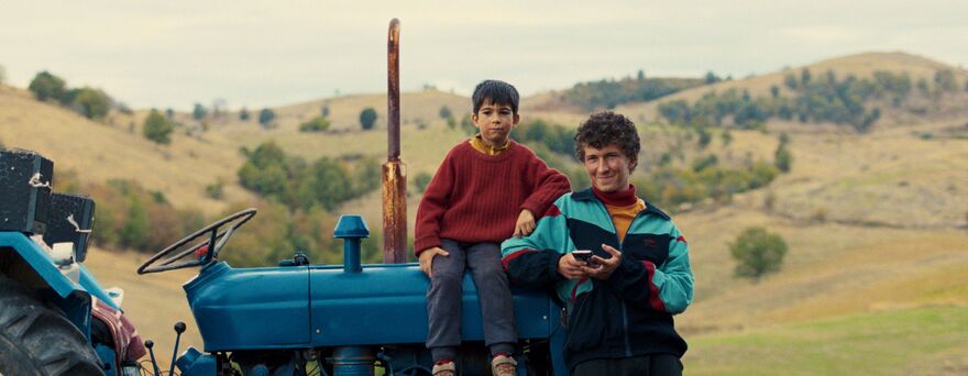 A young teenager leans against a blue tractor as a younger boy sits on it. 
