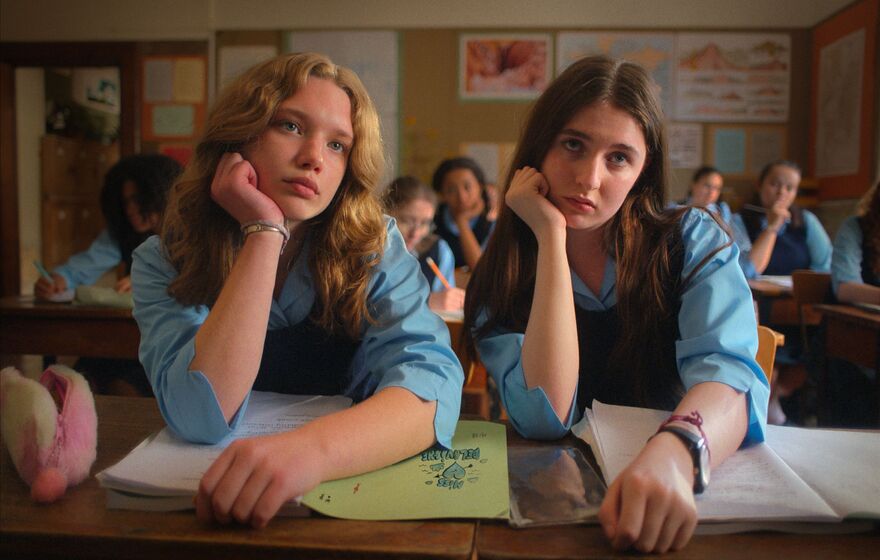 Two teenage girls sit in classroom together, their heads resting on their hands. 