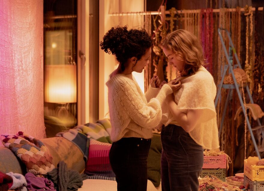 Two women stand close together in a pink toned sitting room.