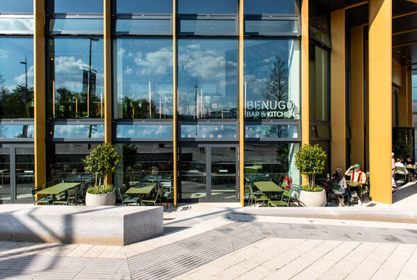 Outside shot of Warwick Arts Centre showing Benugo restaurant sign in the window and outdoor green tables and chairs