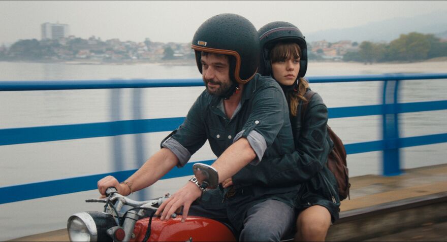 A man and woman ride a motorcycle across a bridge. 