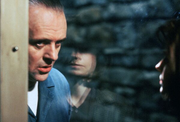 A man stands behind the glass of a locked cell. A woman can be seen in the reflection. 