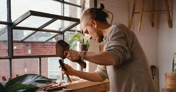 Woodworker Tom Jennings shaping timber at a workbench using a wooden mallet and chisel beside a workshop window.