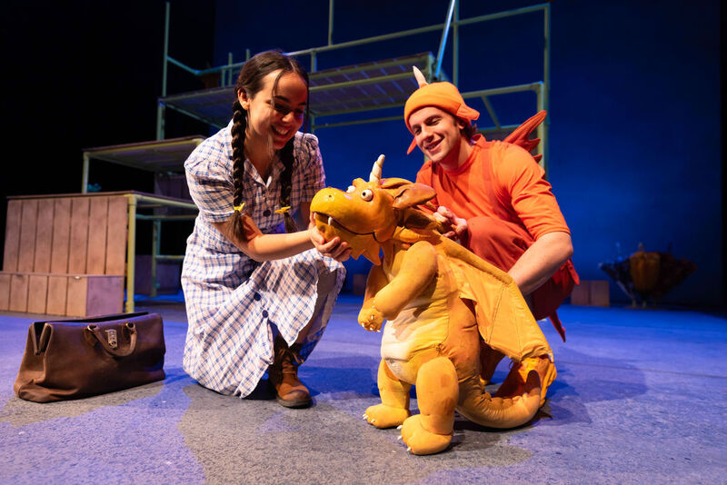 Two smiling actors from the Zog theatre production kneel on stage to playfully interact with a large orange dragon puppet.