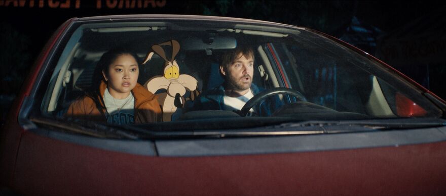 A man and woman sit in a red car with shocked expressions. Wile E. Coyote sits in the back seat. 