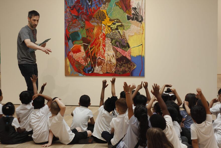 Teacher standing next to an artwork with children sitting on the floor and with their hands up