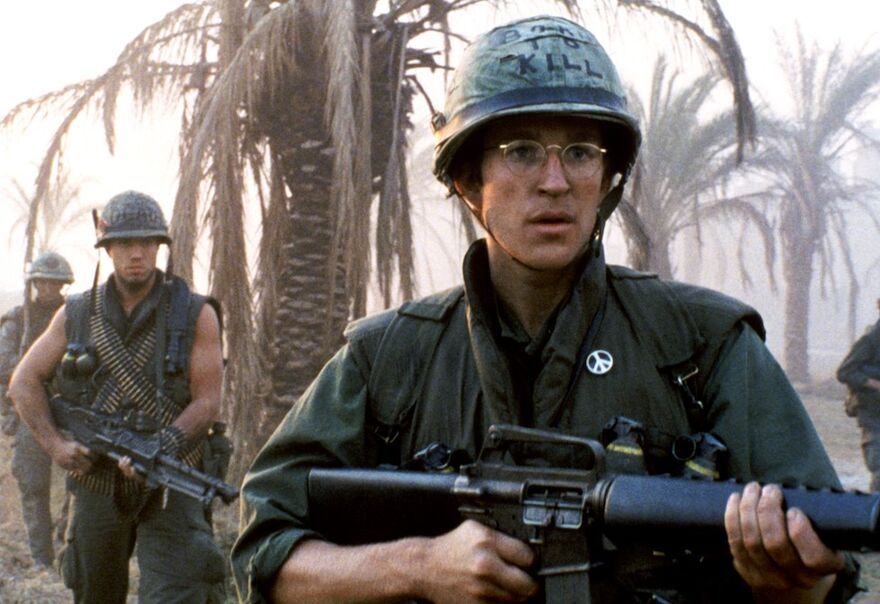 A marine holding a gun. More marines can be seen in the background walking through palm trees. 