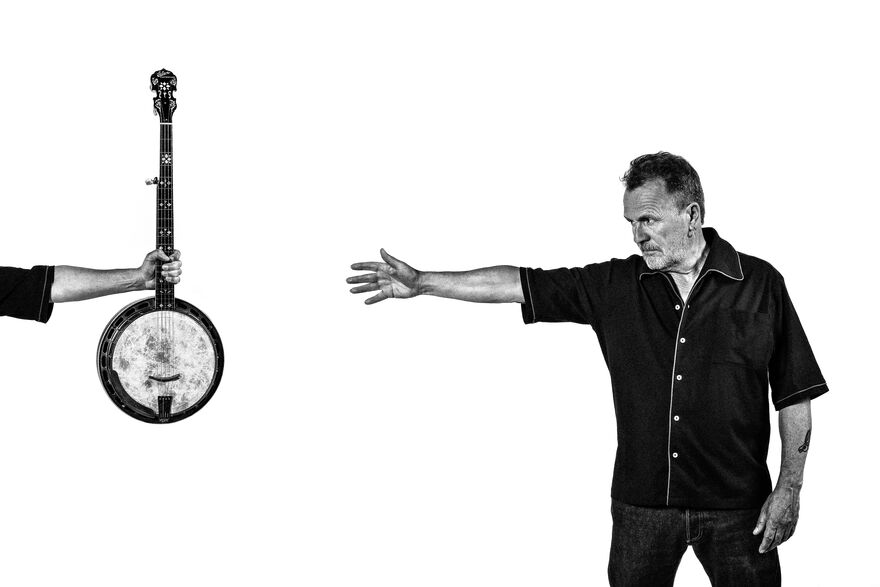 A stylised black and white photo of Martin Simpson reaching for a banjo held by an unseen person against a white background.