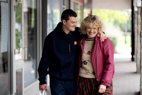 A young man walks with his arm around an older woman. They ae both smiling widely. 