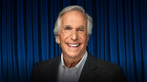 Henry Winkler smiling into the camera in front of a blue curtain