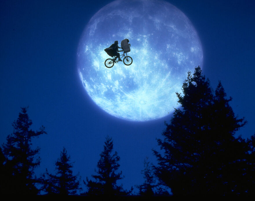 A boy on a flying bike with an alien in the basket covered by a blanket. 