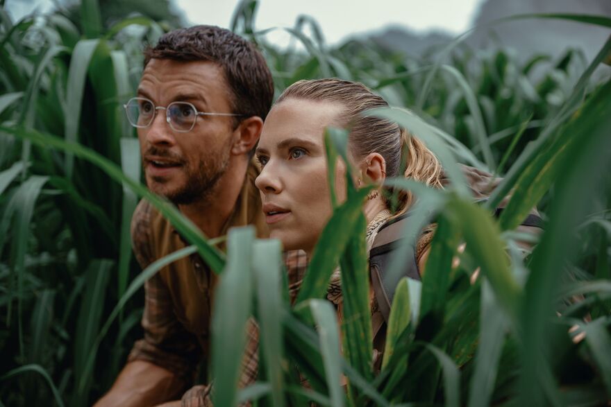 A man and woman hide in long grass looking up at something with marvel.