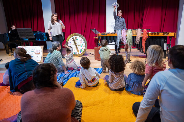 Children and adults sitting on the ground watching two performers play musical instruments.