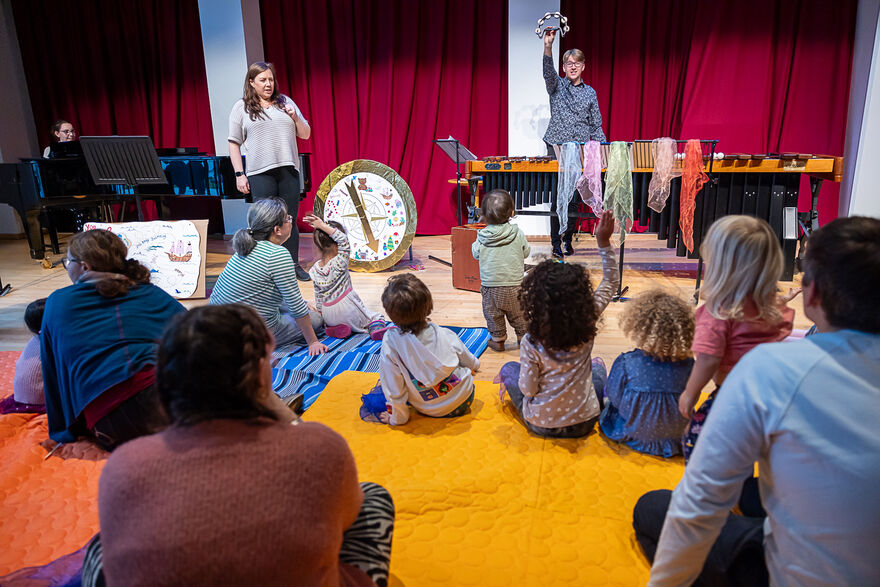 Children and adults sitting on the ground watching two performers play musical instruments.